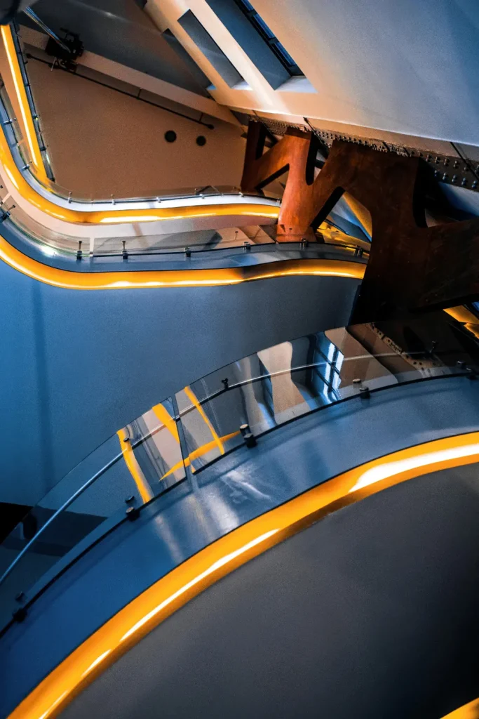 Blue interior space with curved glass railings and glowing yellow-orange LED strips outlining the steps, viewed from a tilted perspective.