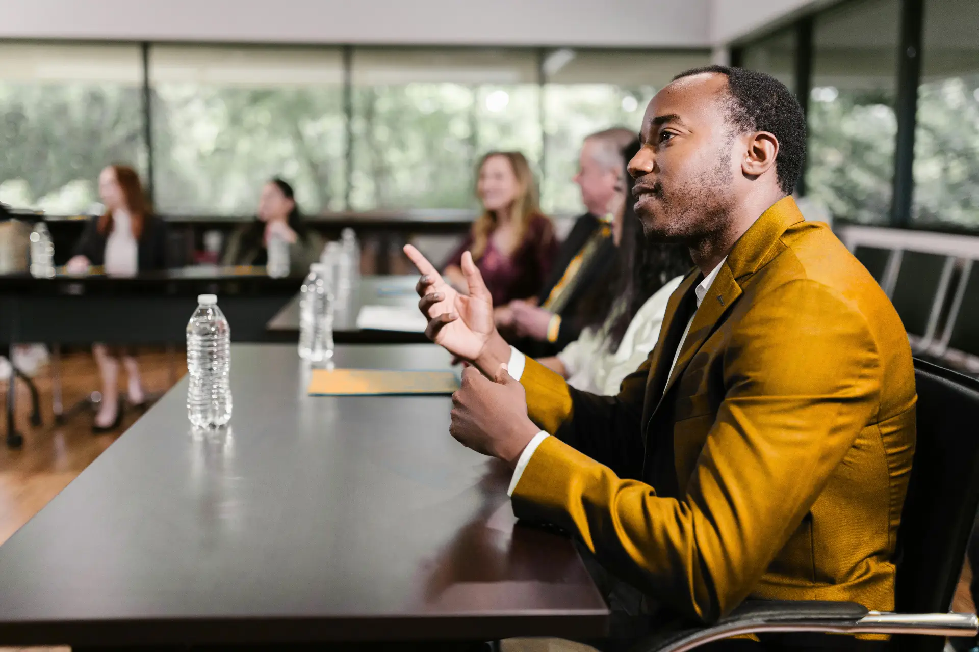 A man in a mustard-yellow blazer sits at a long conference table, speaking with hand gestures. In the background, several blurred colleagues sit and listen, with water bottles and folders on the table and large windows showing greenery outside.
