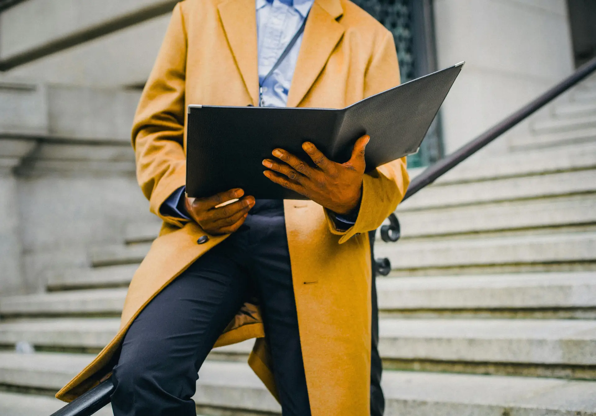 Professional on the Go with Notebook Person wearing a camel-colored coat and blue shirt sits on outdoor stairs, holding an open black folder or binder with both hands.