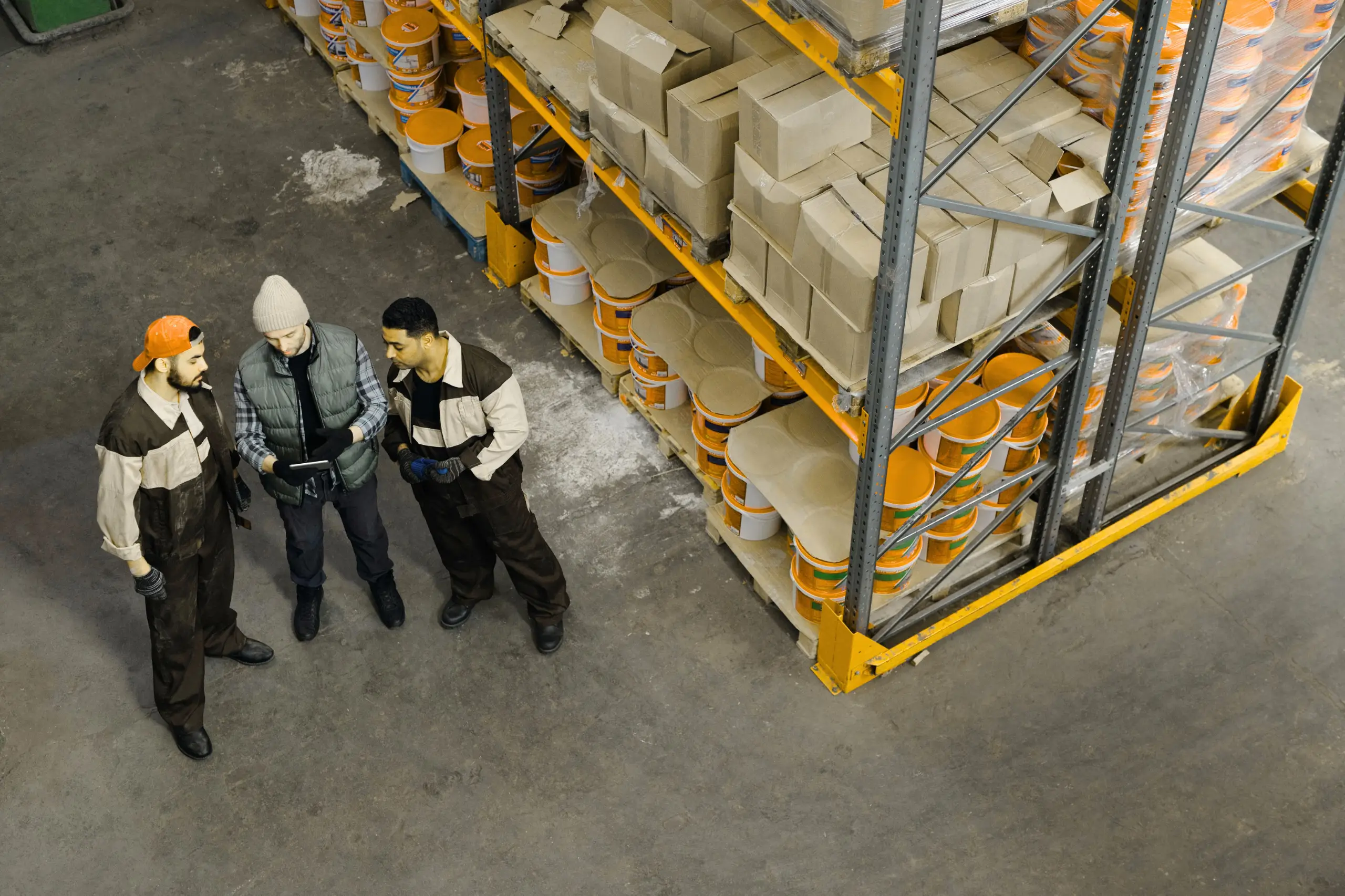 Three workers stand in a warehouse aisle looking at a tablet. They wear work overalls, with one in an orange cap and another in a gray beanie, surrounded by tall orange-yellow metal shelving with boxes and paint cans.