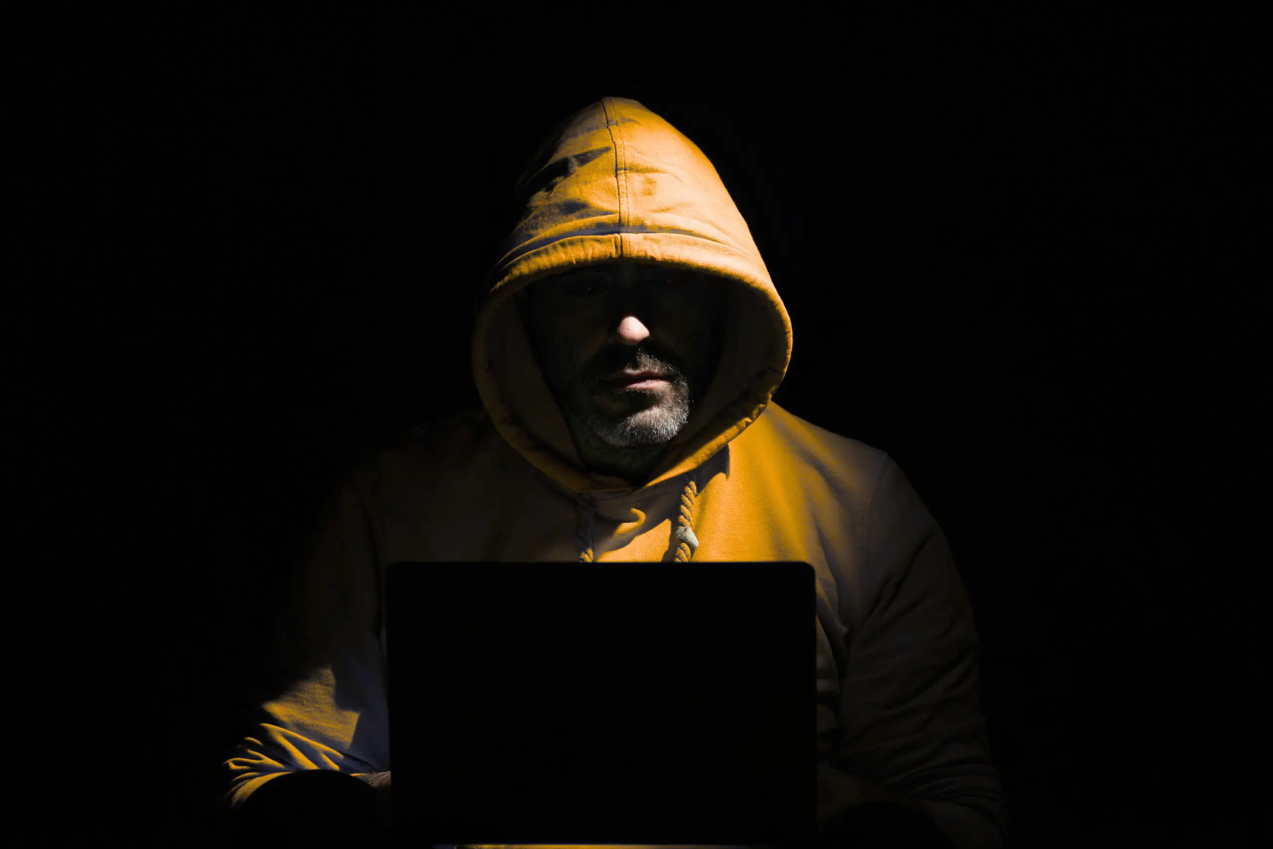 A person wearing a yellow hoodie sits in darkness, their face partially lit by the glow of a laptop screen.