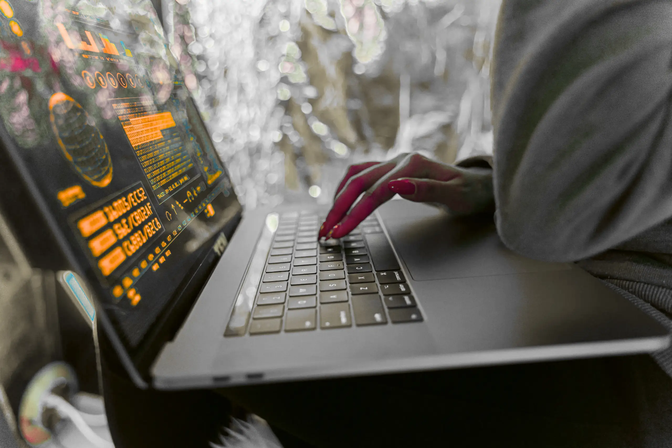 Close-up of a person typing on a laptop with an orange-tinted code display on the screen. The scene has a blurred metallic background and the person's red-painted fingernails on the keyboard.