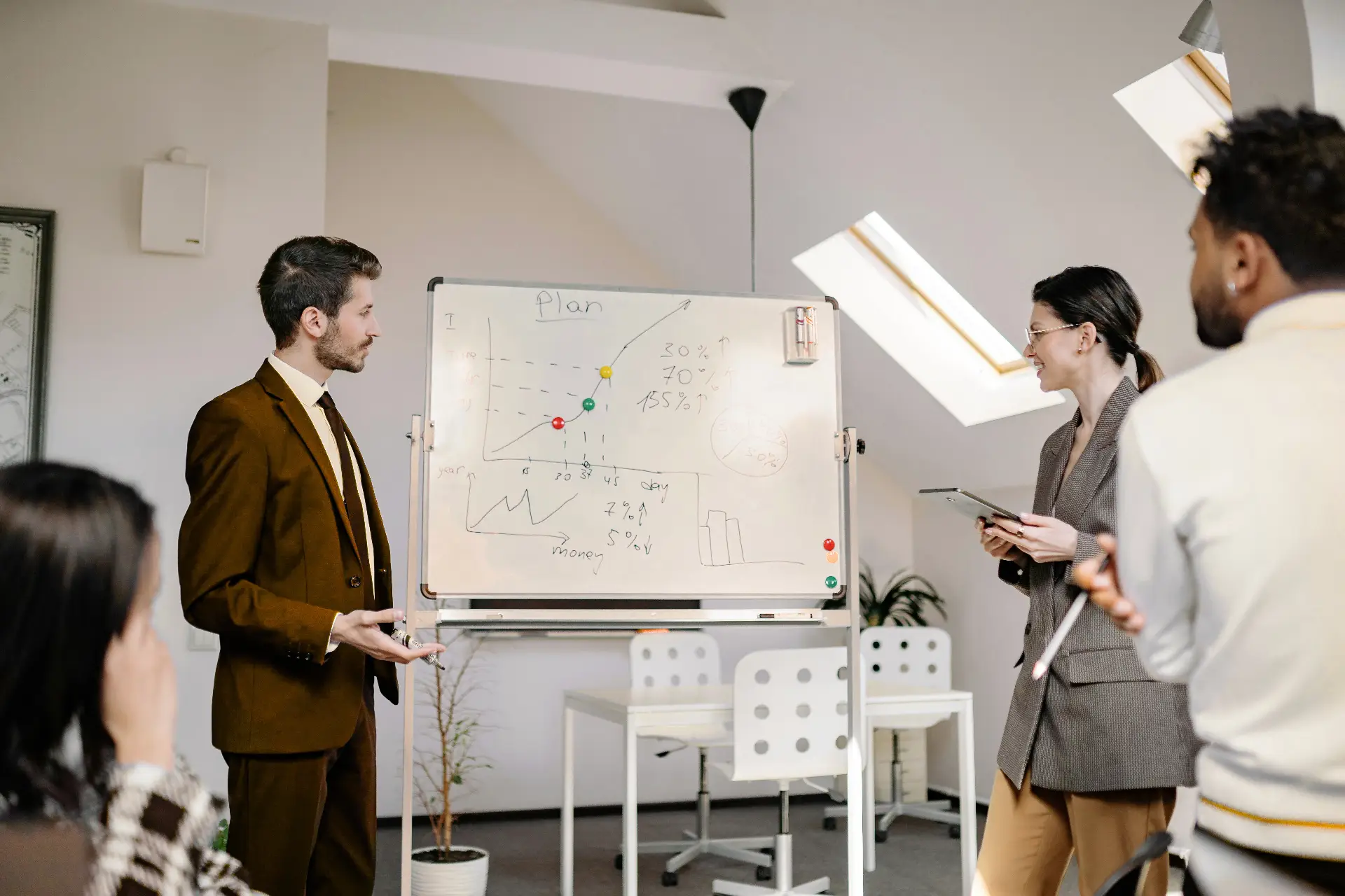 A man in a brown suit presents beside a whiteboard with charts, while a woman in a gray blazer holds a tablet and smiles. Two colleagues watch from the foreground in a bright office space with modern furniture.
