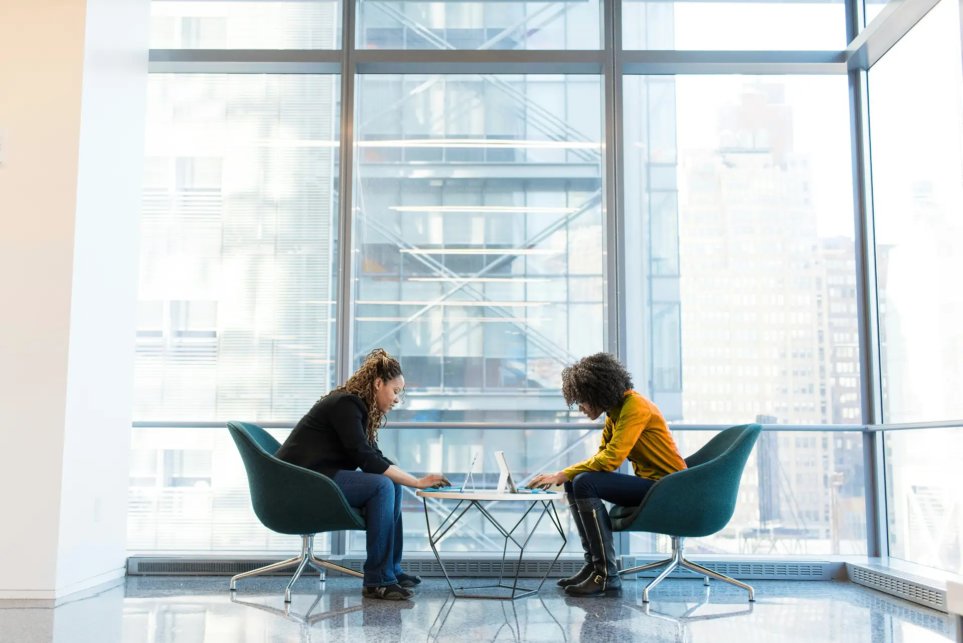 Two people sit in teal swivel chairs at a small glass table, each focused on their laptops. They face each other in a bright, open office with large floor-to-ceiling windows and a cityscape outside.