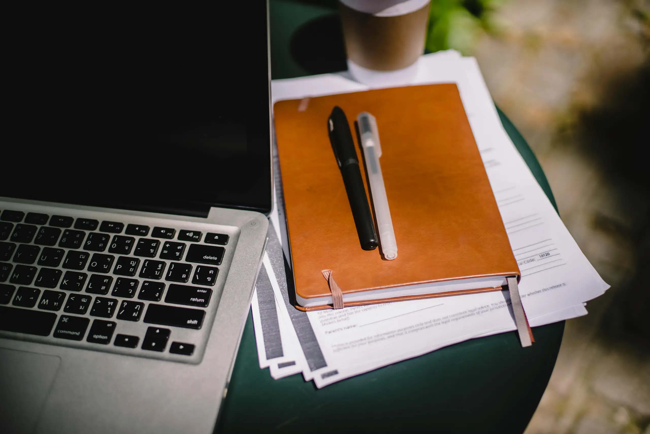 Close-up of a silver laptop keyboard on the left, with an orange-brown notebook and two pens resting on a stack of papers on a green table outdoors.