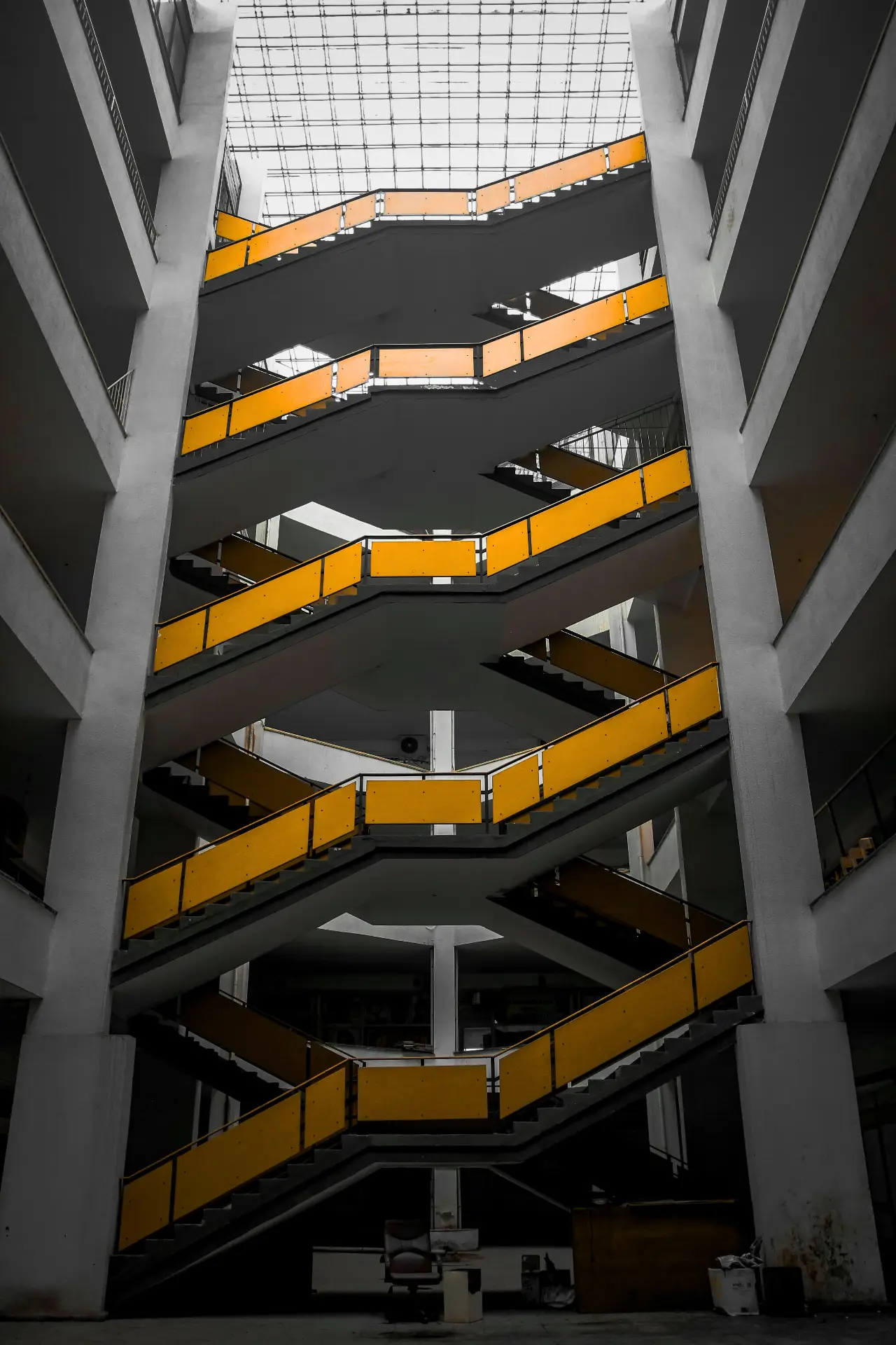 Vertical shot of a multi-level interior atrium with zigzagging yellow handrails on dark gray stairs, framed by tall concrete columns and a skylight above.