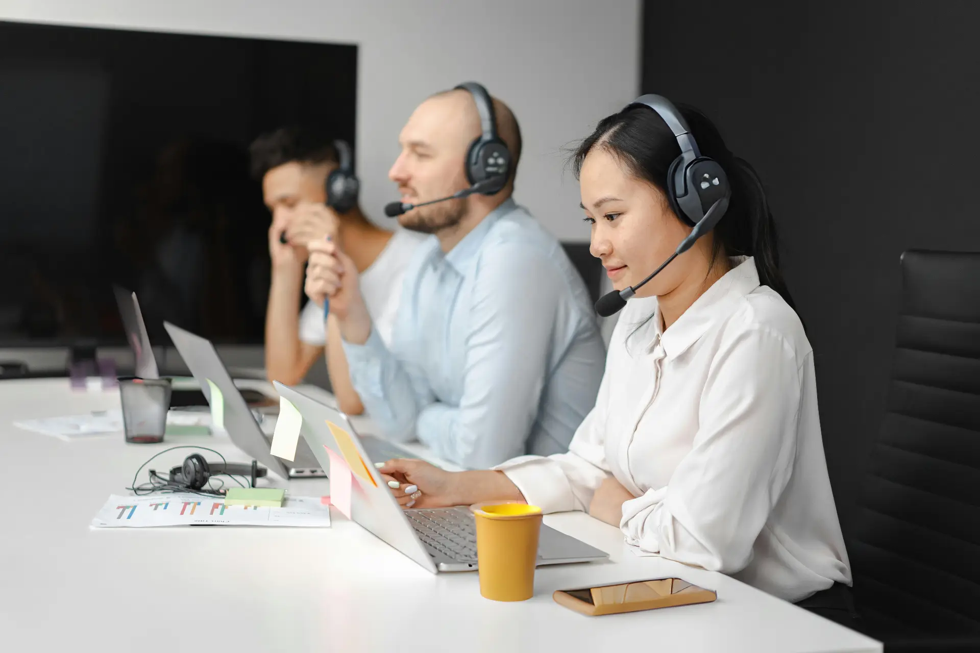 Three agents wearing headsets sit at a white desk with laptops, notes, and a yellow coffee cup. A large screen in the background and a focused female agent typing on her laptop.