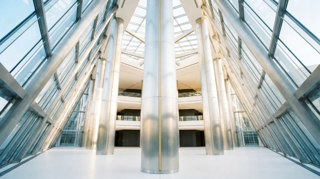 Low-angle view inside a modern building atrium with tall cylindrical white columns and curved walls of blue-tinted glass windows on both sides, looking up toward a bright skylight ceiling.