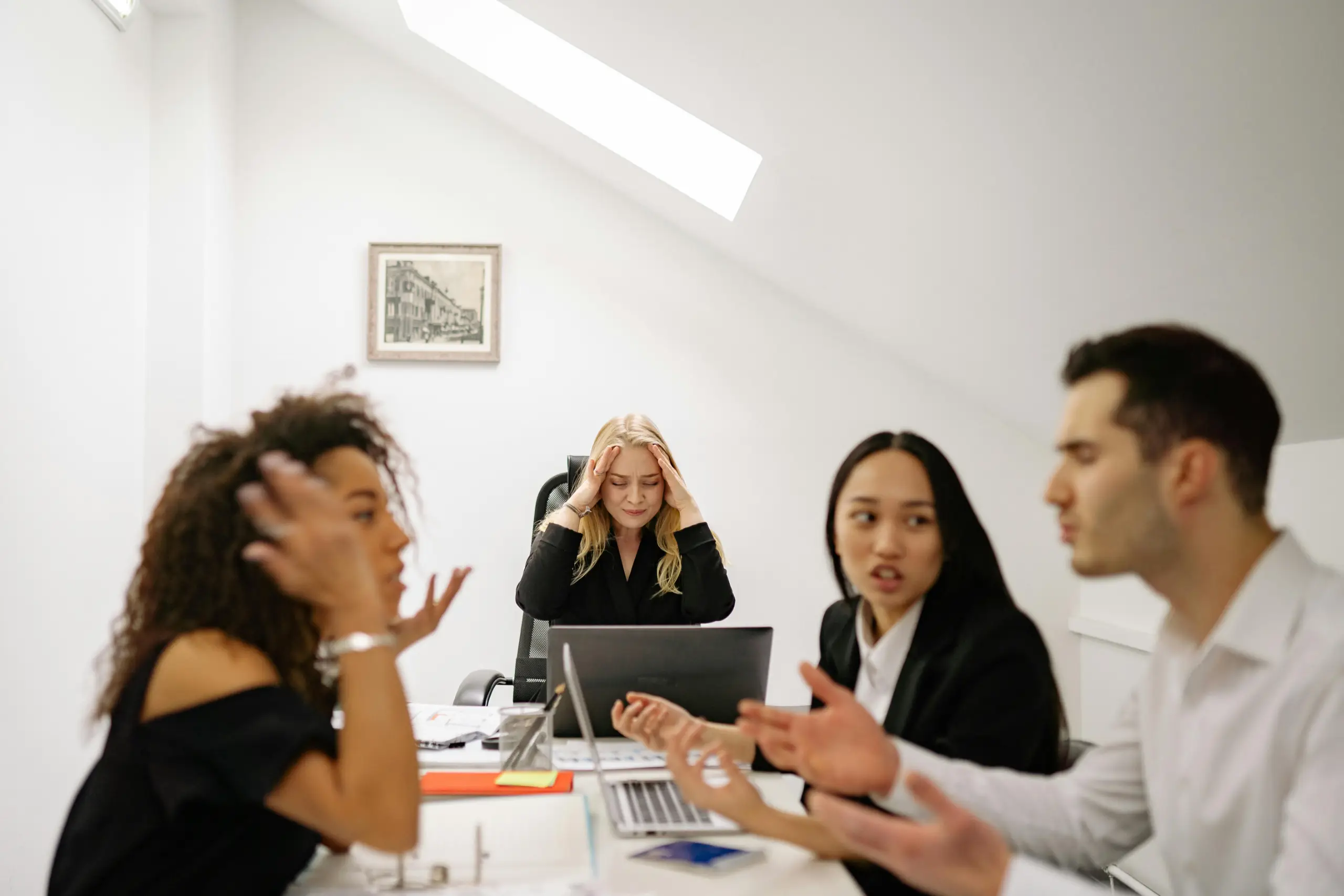 Four coworkers sit around a conference table in a bright office. A blonde woman in the center holds her head in frustration while the others gesture and talk. A laptop and documents are on the table, with a framed picture on the white wall behind.