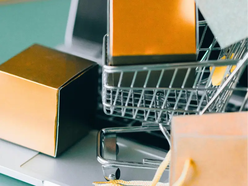 Metal shopping cart filled with orange and brown cardboard boxes placed on a shelf, with more boxes and packaging material nearby.