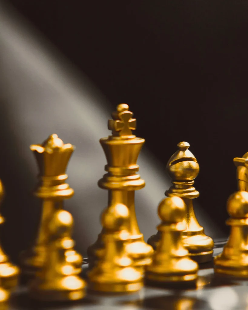 Shiny gold chess pieces, including a central king-like piece, with several pawns and rooks around it. The scene is softly lit with a blurred dark background and shallow depth of field.