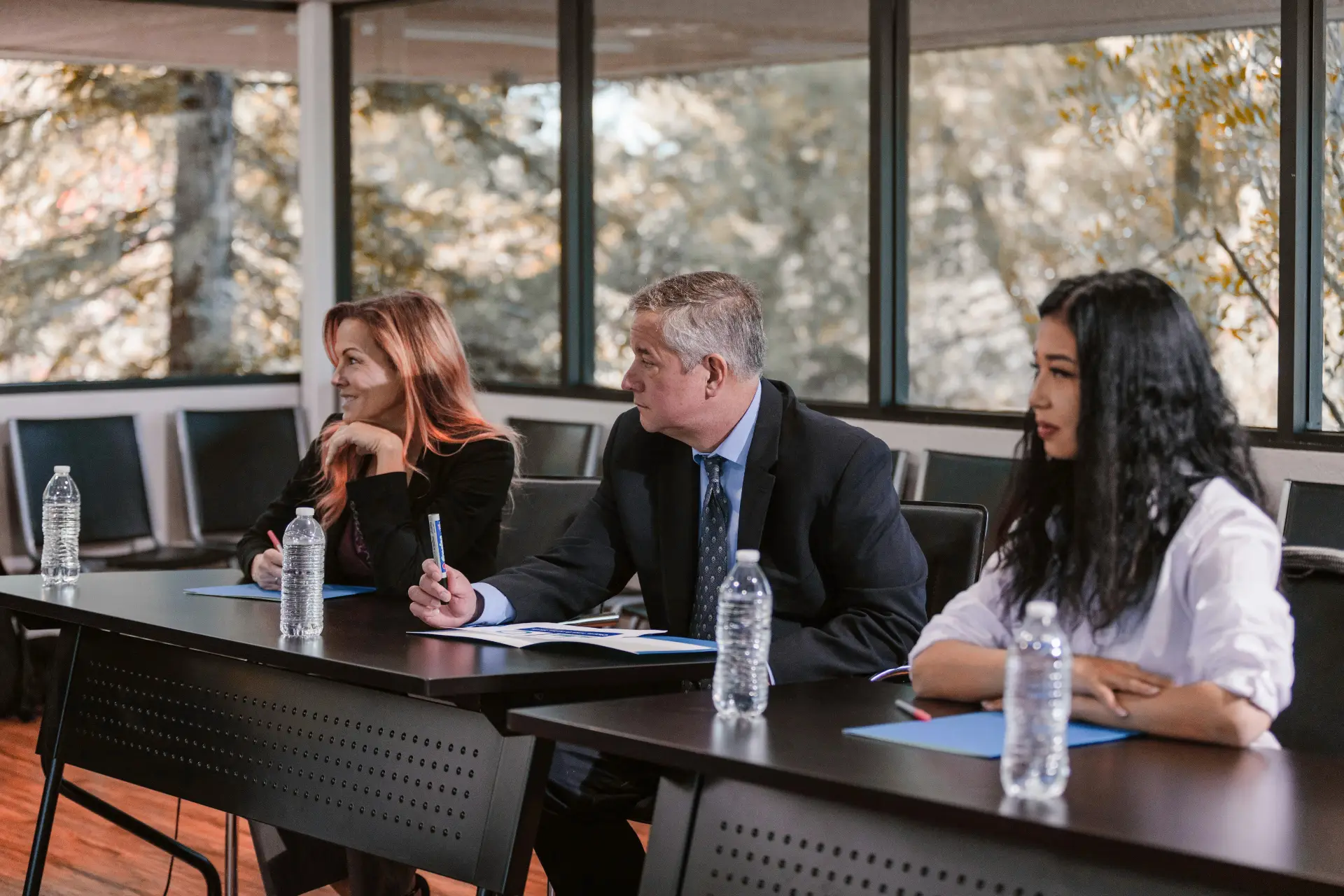 Three professionals seated at a dark conference table with notebooks and water bottles. A man in a suit center foreground, flanked by a woman with pink hair on the left and a woman with long dark hair on the right, large windows and autumn trees in the background.