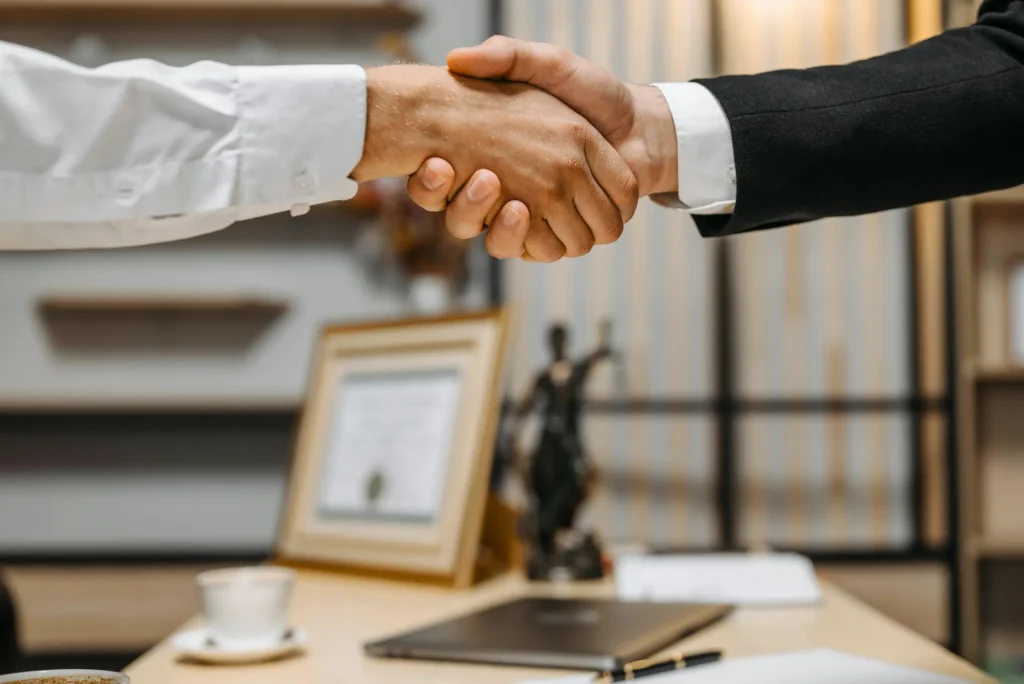 Two business professionals shake hands across an office desk, with computers and documents blurred in the background, reflecting align it spend with ebitda priorities.