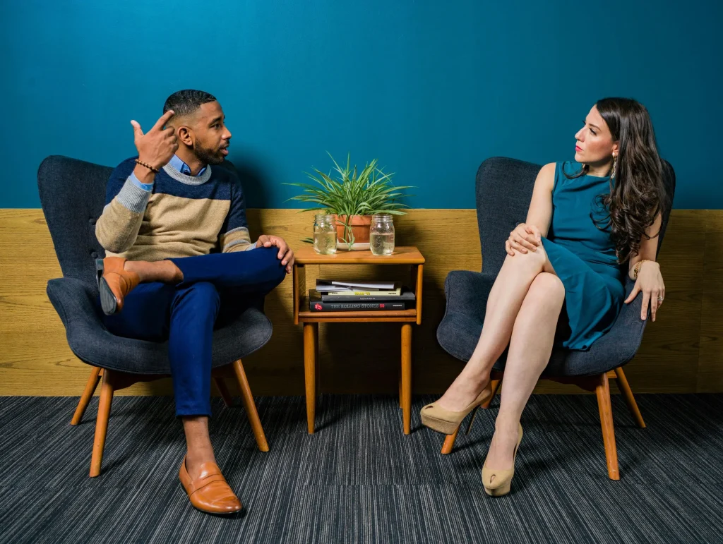 Two business professionals sit facing each other on chairs in a modern meeting room with a small table holding a potted plant and books, discussing digital transformation consulting.