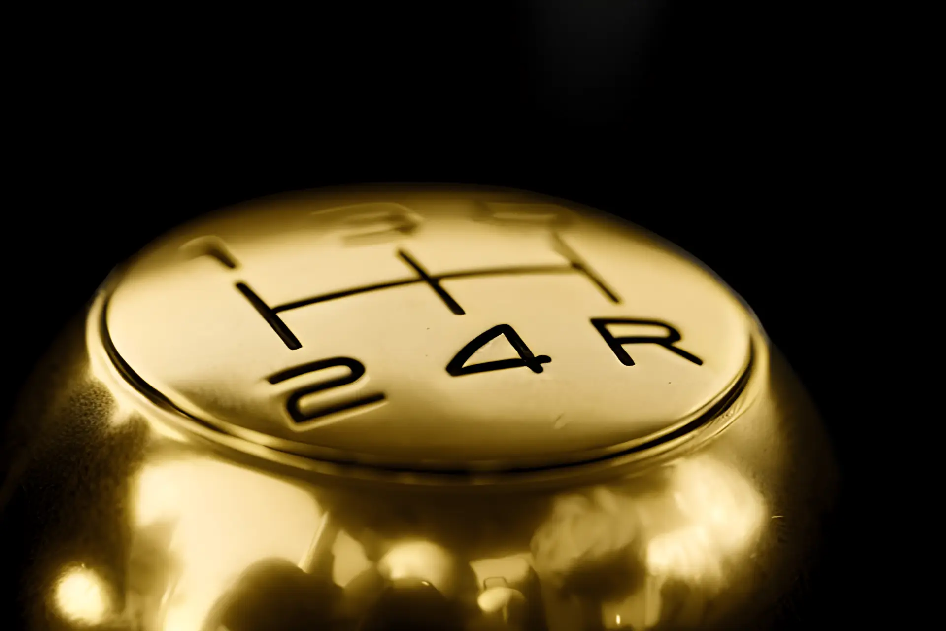 Close-up of a gold-tinted manual car gear knob showing the shift pattern with positions for first through fifth gears and reverse, against a dark background.
