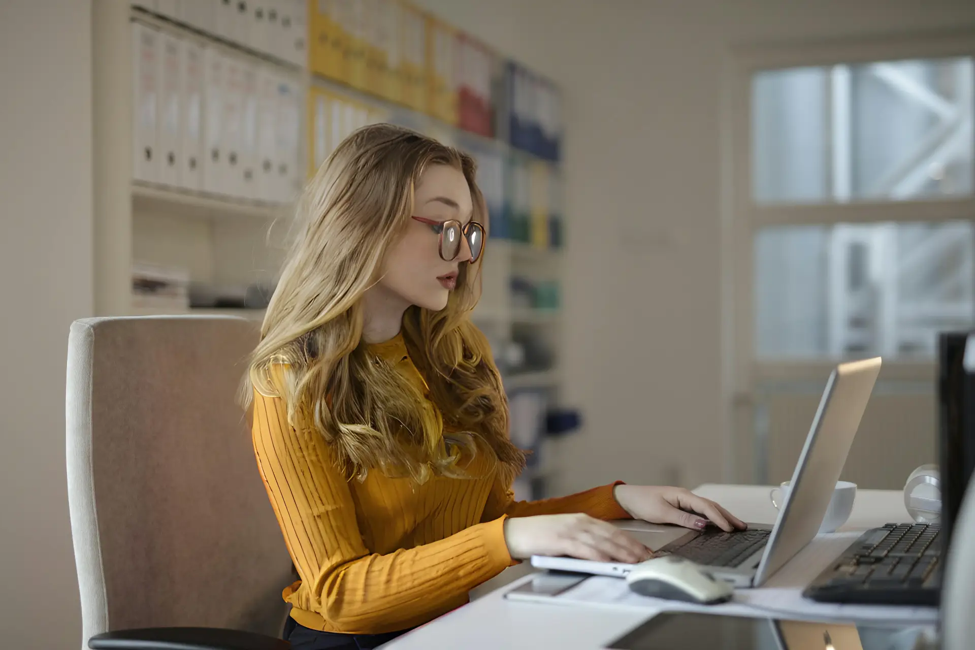 Professional woman with long blond hair and glasses sits at a desk in an office, typing on a silver laptop. She wears a mustard yellow ribbed top, and there are organized shelves with binders in the background.