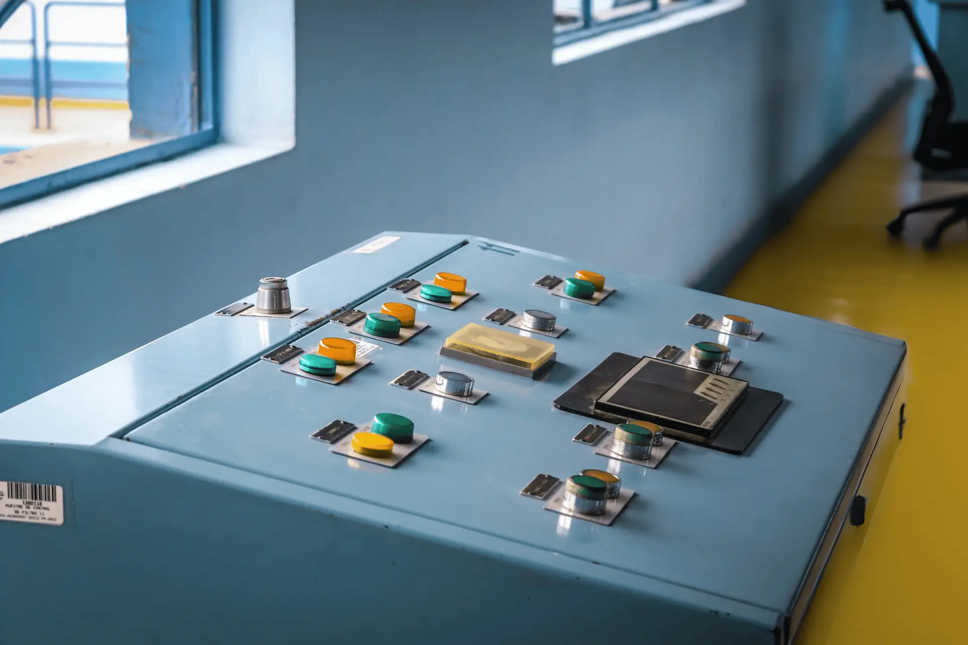 A blue industrial control panel with multiple green and yellow buttons and a few silver toggles, located in a room with a window and yellow floor trim.