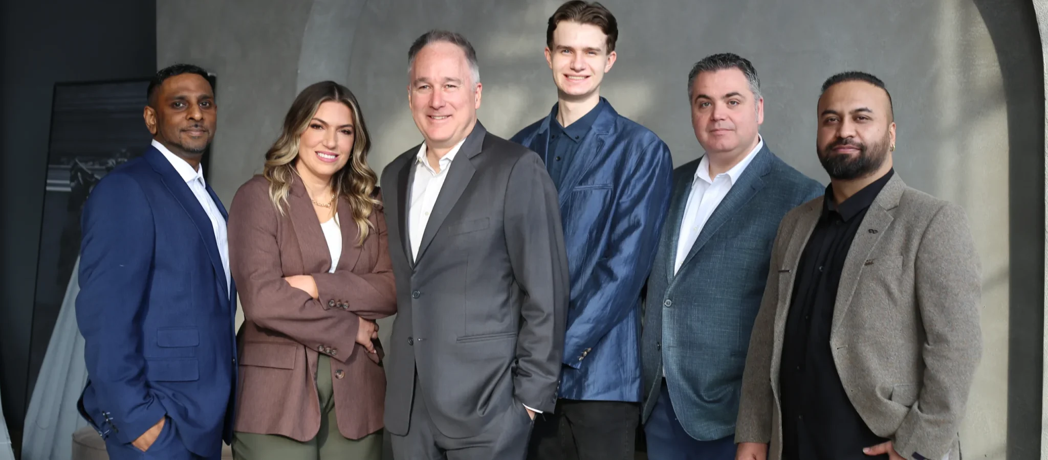 Group of six professionals in blue, gray, and brown suits pose in a modern office setting, showcasing teamwork and business leadership. The Narrative Group.
